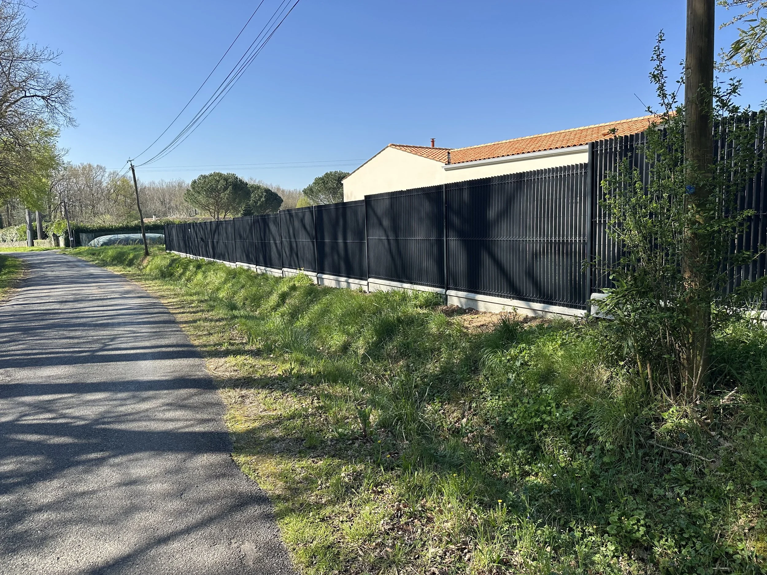 Une clôture noire longe une route rurale avec des arbres et un ciel bleu.