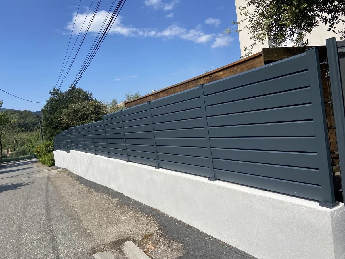 Clôture en métal gris installée le long d'un mur blanc, avec une route et un ciel bleu en arrière-plan.
