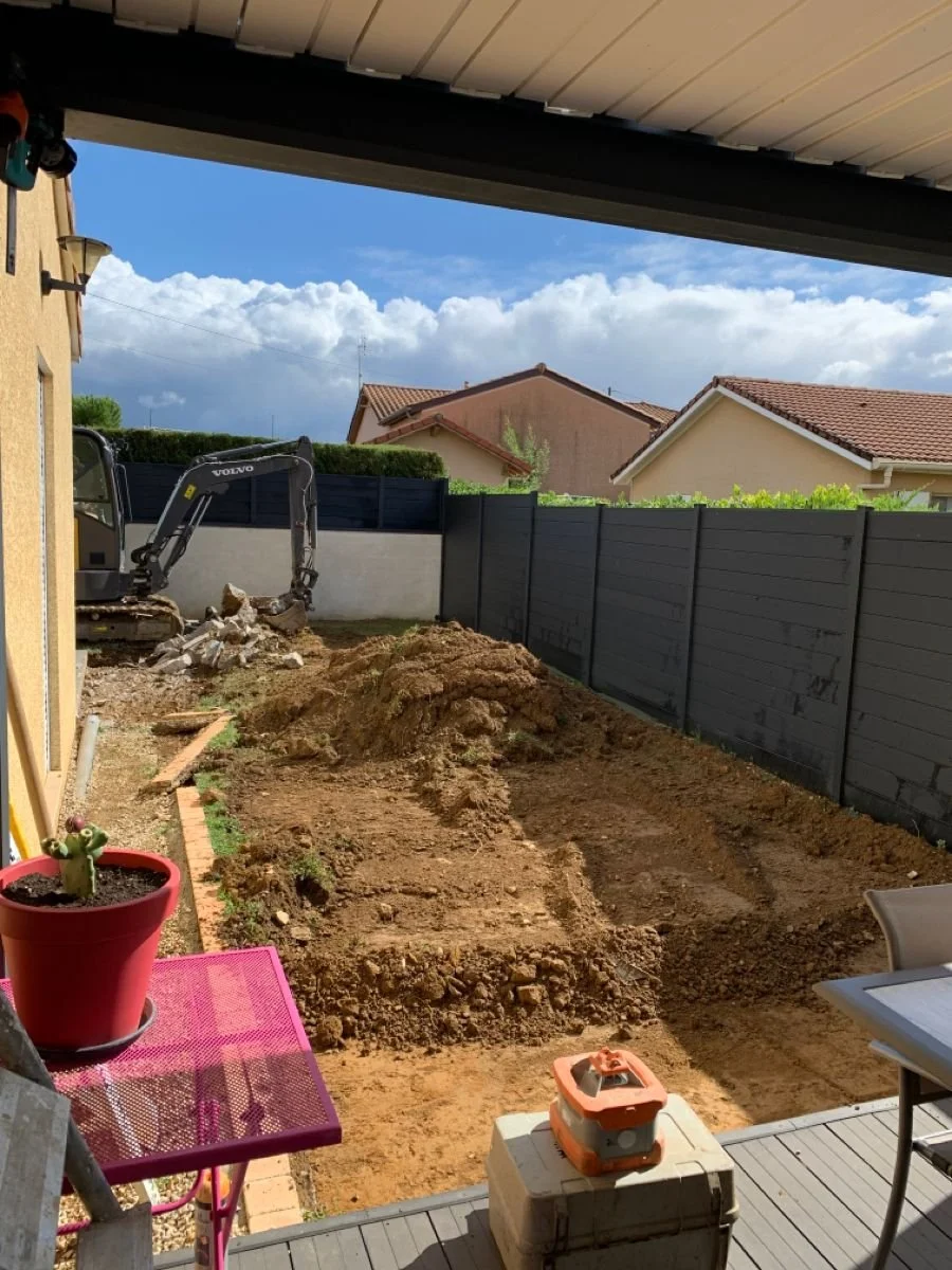 Travaux de terrassement dans un jardin, avec une pelle mécanique et du sol en cours de déplacement, vue depuis une terrasse couverte