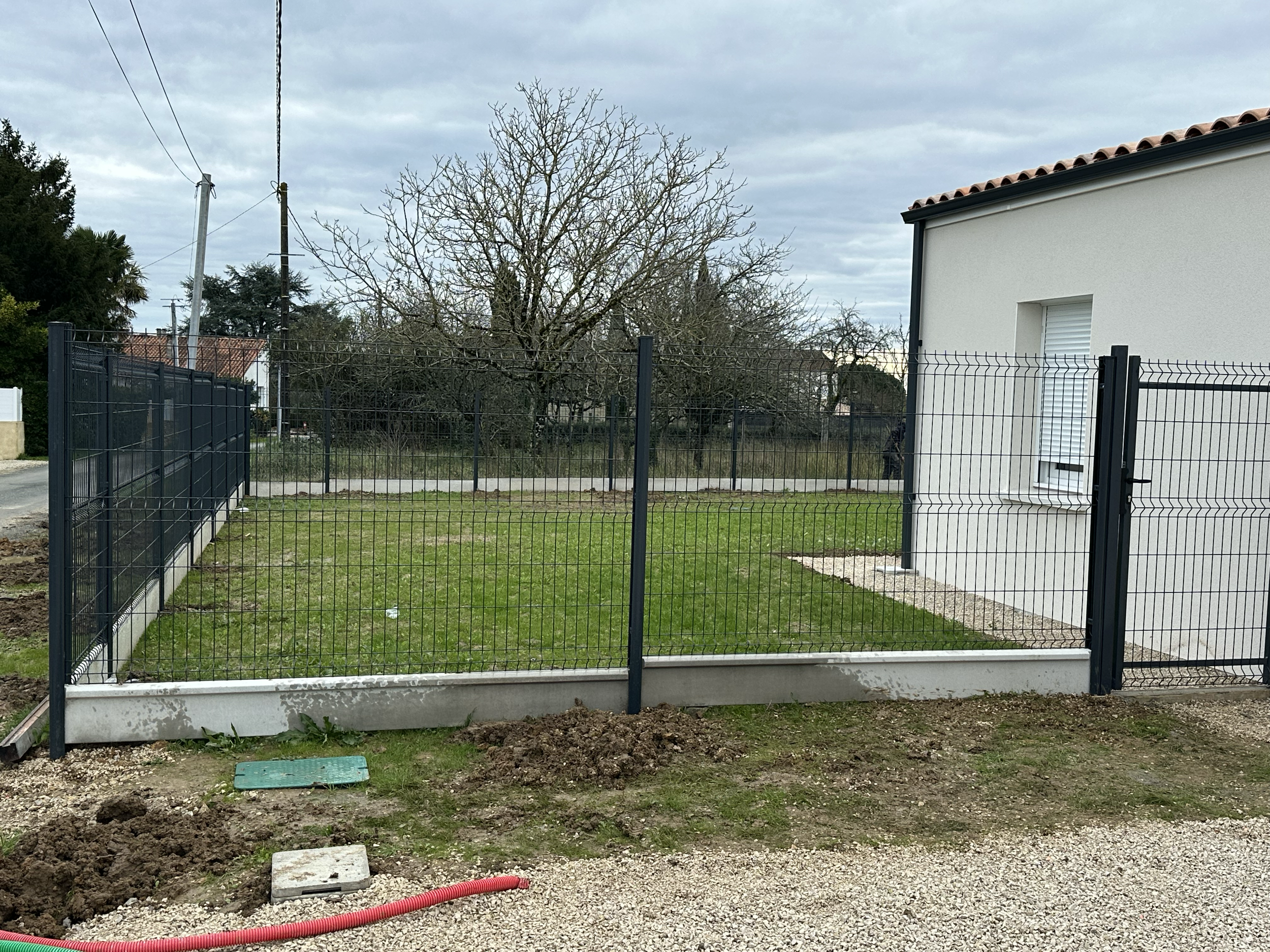Un jardin clôturé avec une pelouse verte, une maison blanche à droite, un arbre sans feuilles, et un ciel nuageux.