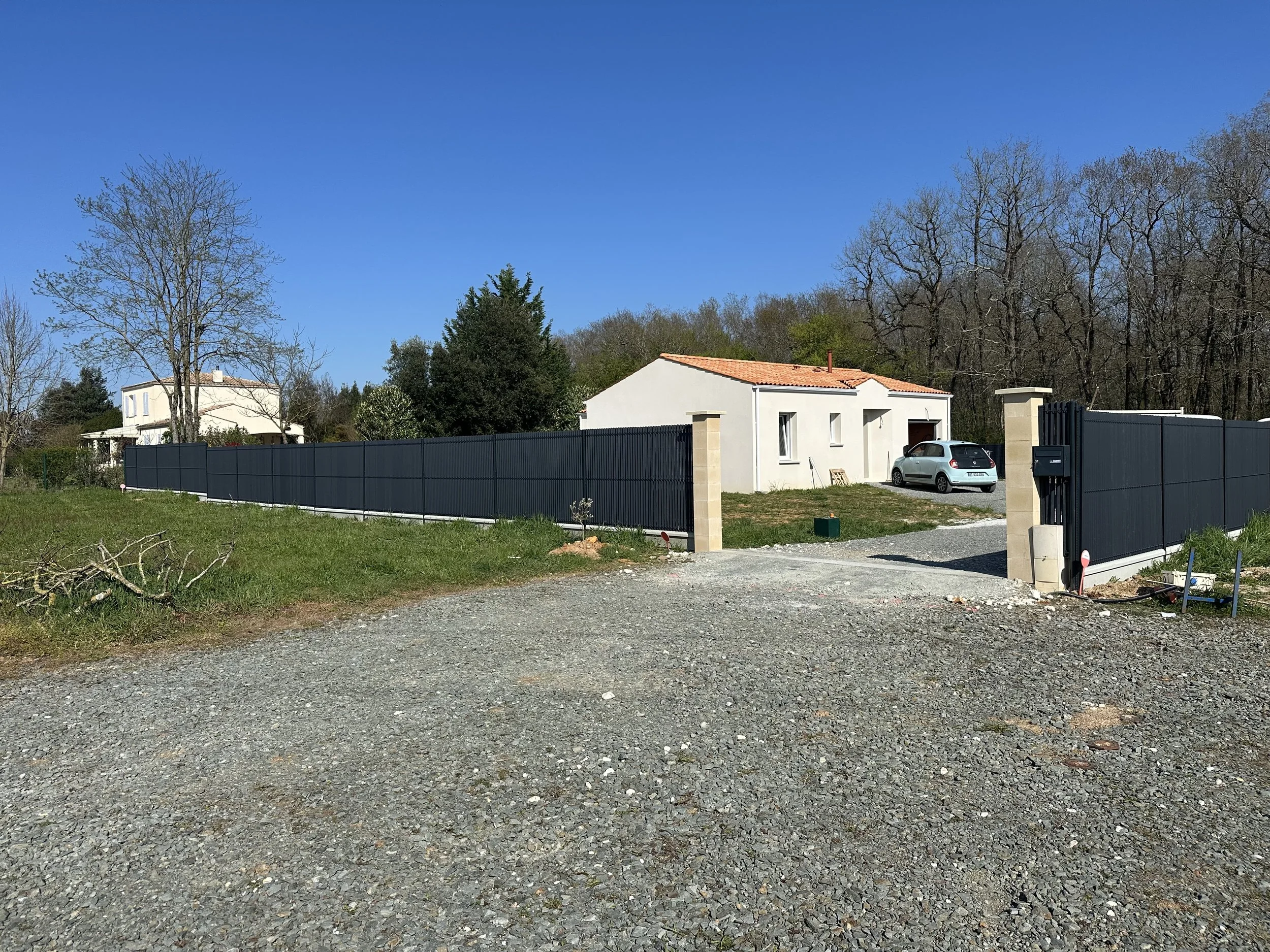 Une maison récente avec un mur en clôture noire, une voiture blanche dans l'allée, entourée d'arbres et d’un ciel bleu clair.