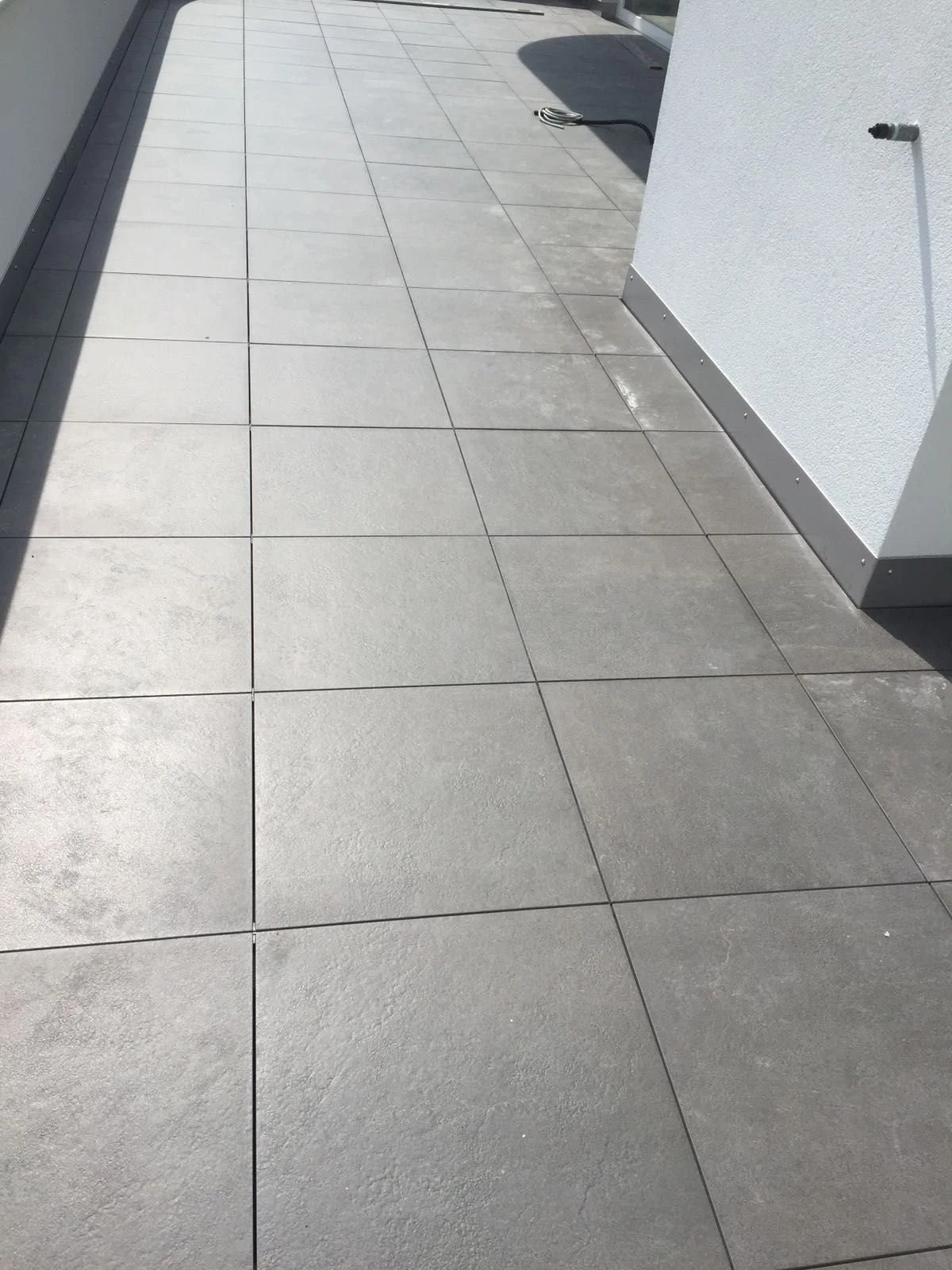 Terrasse en béton avec de larges dalles de sol, bordée d'un mur blanc et d'une façade en verre. Un tuyau d'arrosage est visible au fond.