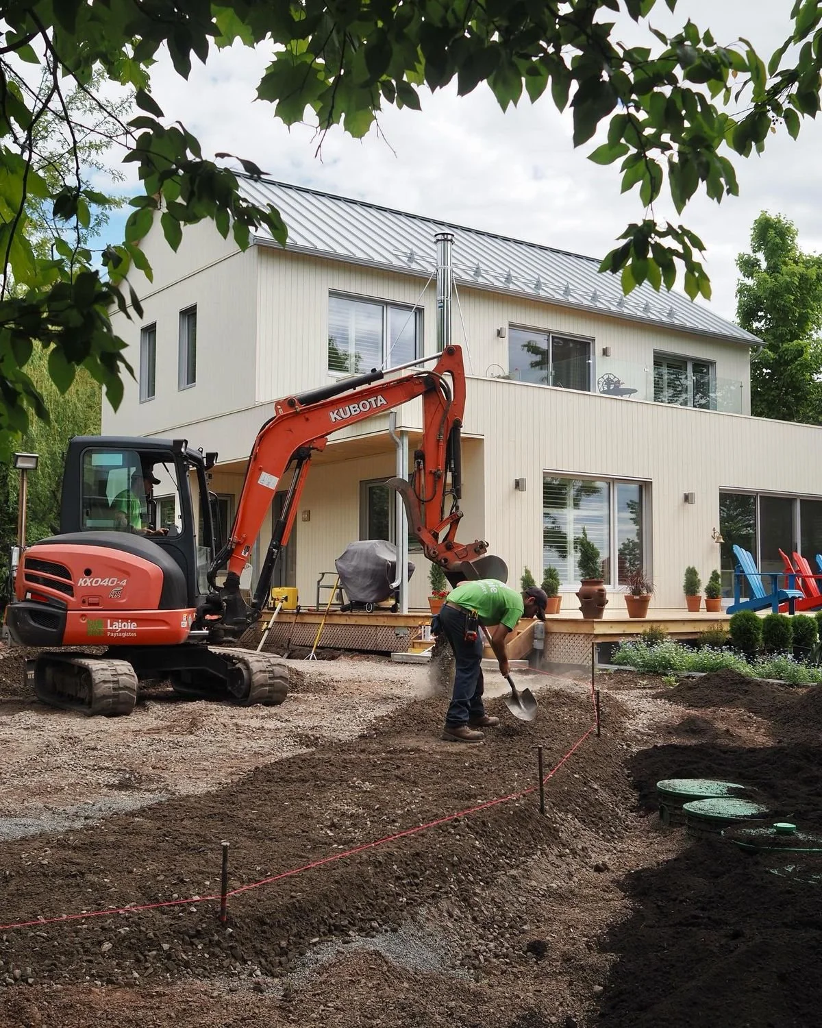 Travail de construction dans le jardin d'une maison moderne, avec une pelleteuse orange, un ouvrier en vert utilisant une pelle, et des pots de fleurs près de la maison.