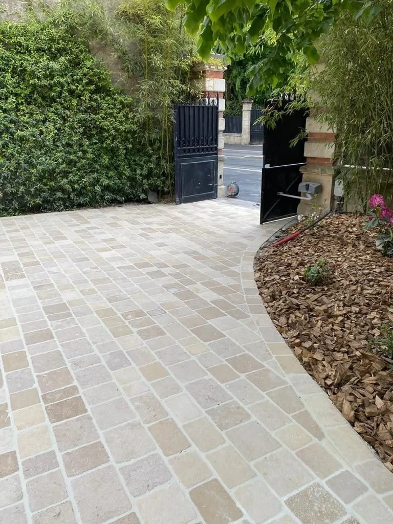 Vue d'une allée pavée avec un portail noir, entourée de végétation et de fleurs, et une bordure en bois mulch.