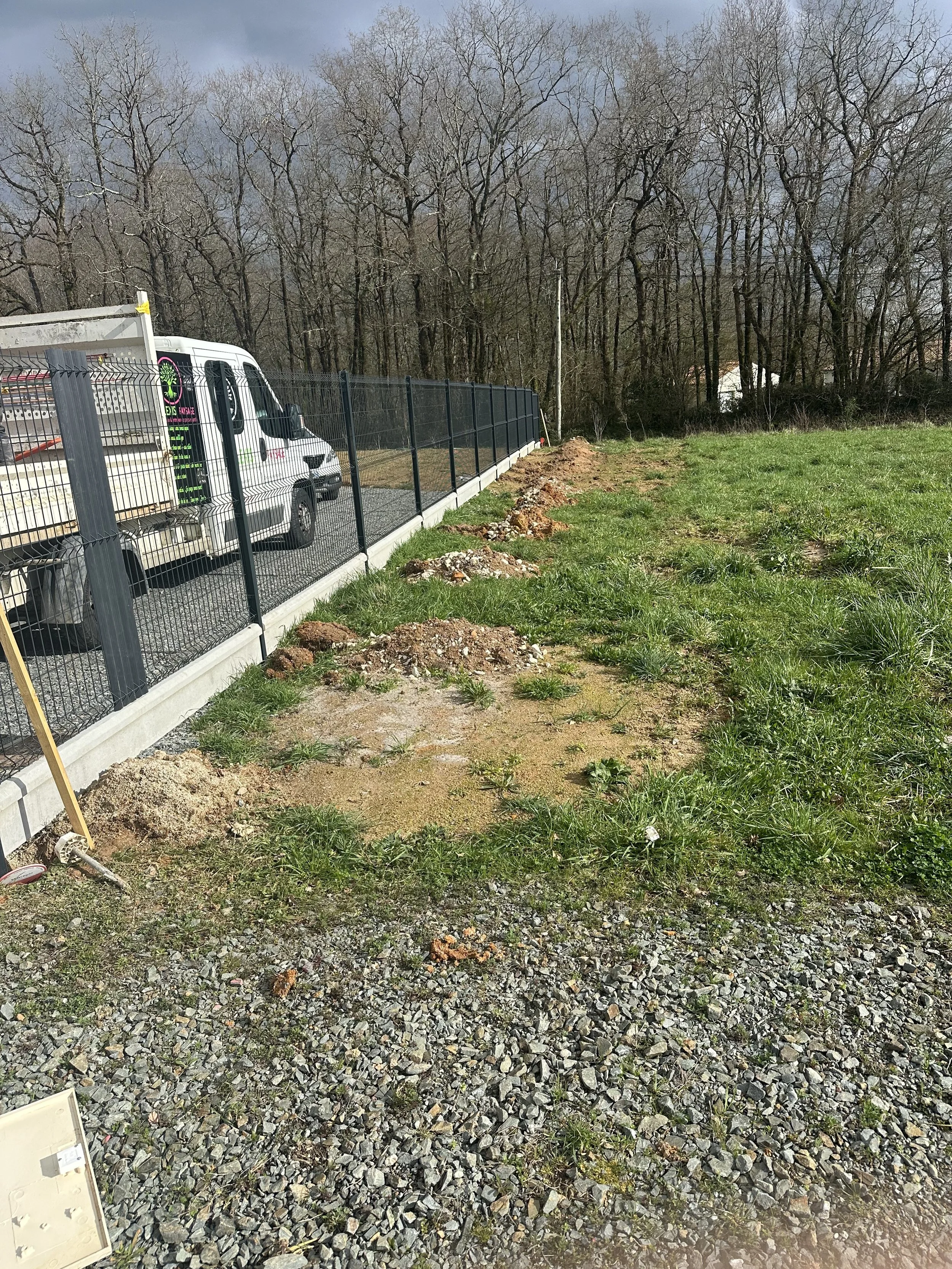 Travaux en cours dans un espace extérieur, avec une clôture en métal noire, une voiture blanche, un arbre sans feuilles et un terrain en terre et herbe.