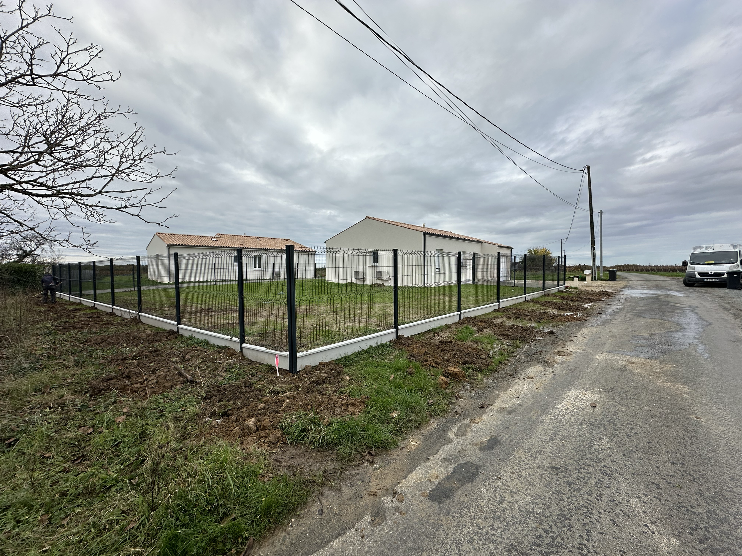 Maisons neuves entourées d'une clôture noire et une route dégradée, ciel nuageux.
