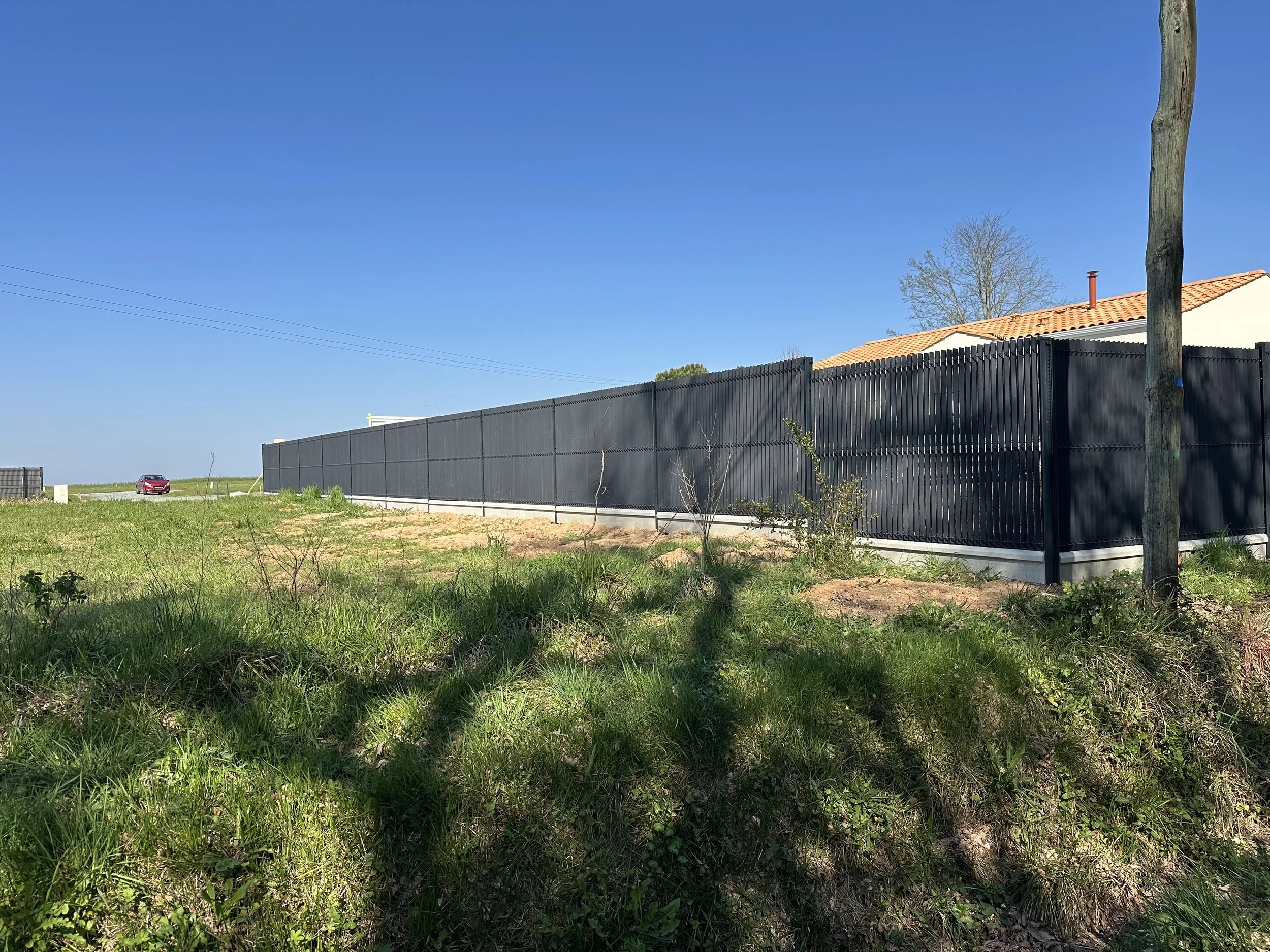 Clôture noire en métal le long d'un bâtiment avec un toit en tuiles orange, sous un ciel bleu clair, avec un arbre et une ligne électrique