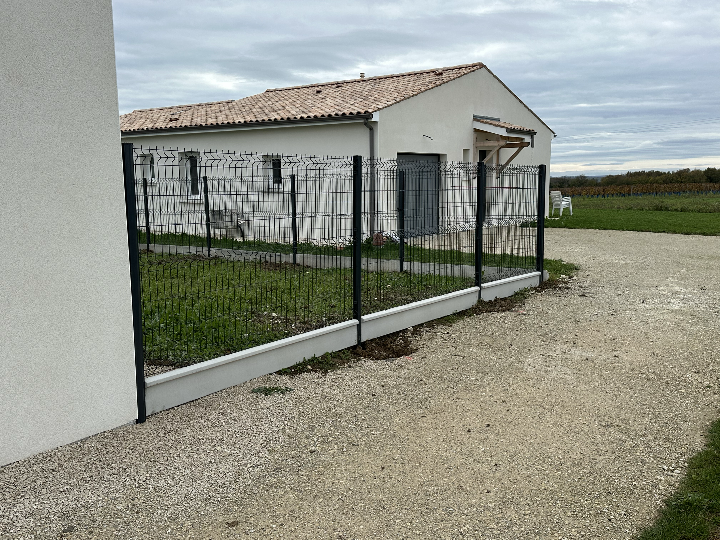 Une clôture métallique noire entourant un petit jardin face à une maison avec un garage, le tout dans un environnement rural avec un ciel nuageux.