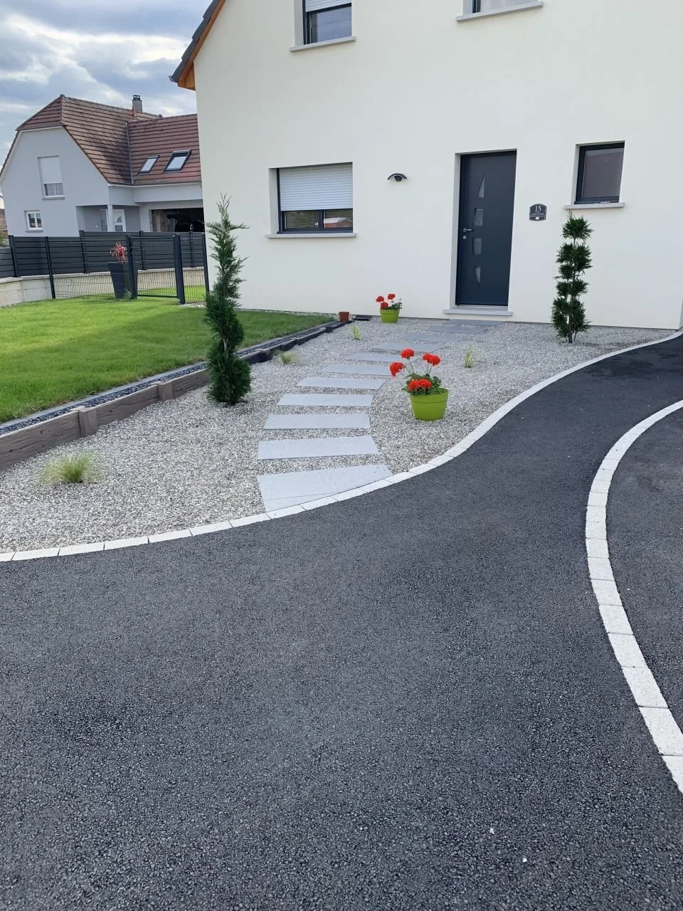 Allée pavée devant une maison moderne blanche avec un jardin bien entretenu, deux petits arbres et des plantes en pot colorées.