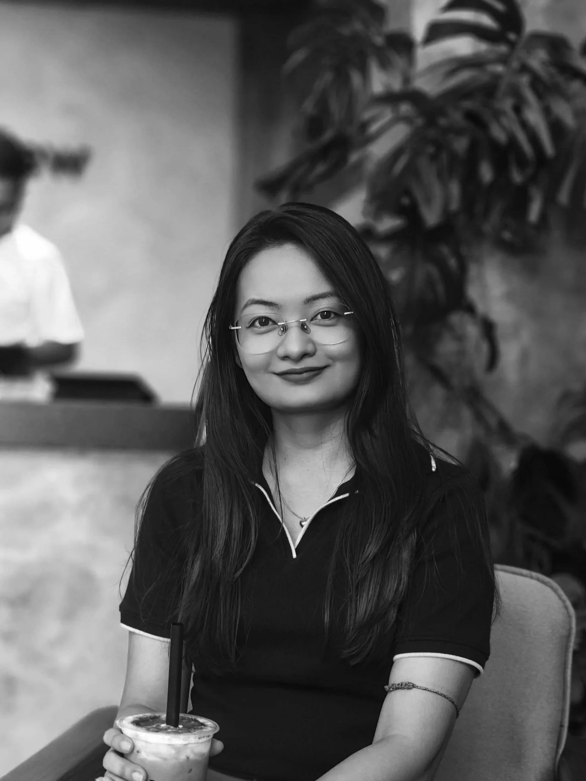 Black and white photograph of a young woman with long dark hair, wearing glasses and a short-sleeved shirt, sitting at a table holding a drink with a straw. There are large plants in the background and a blurred figure in the background.