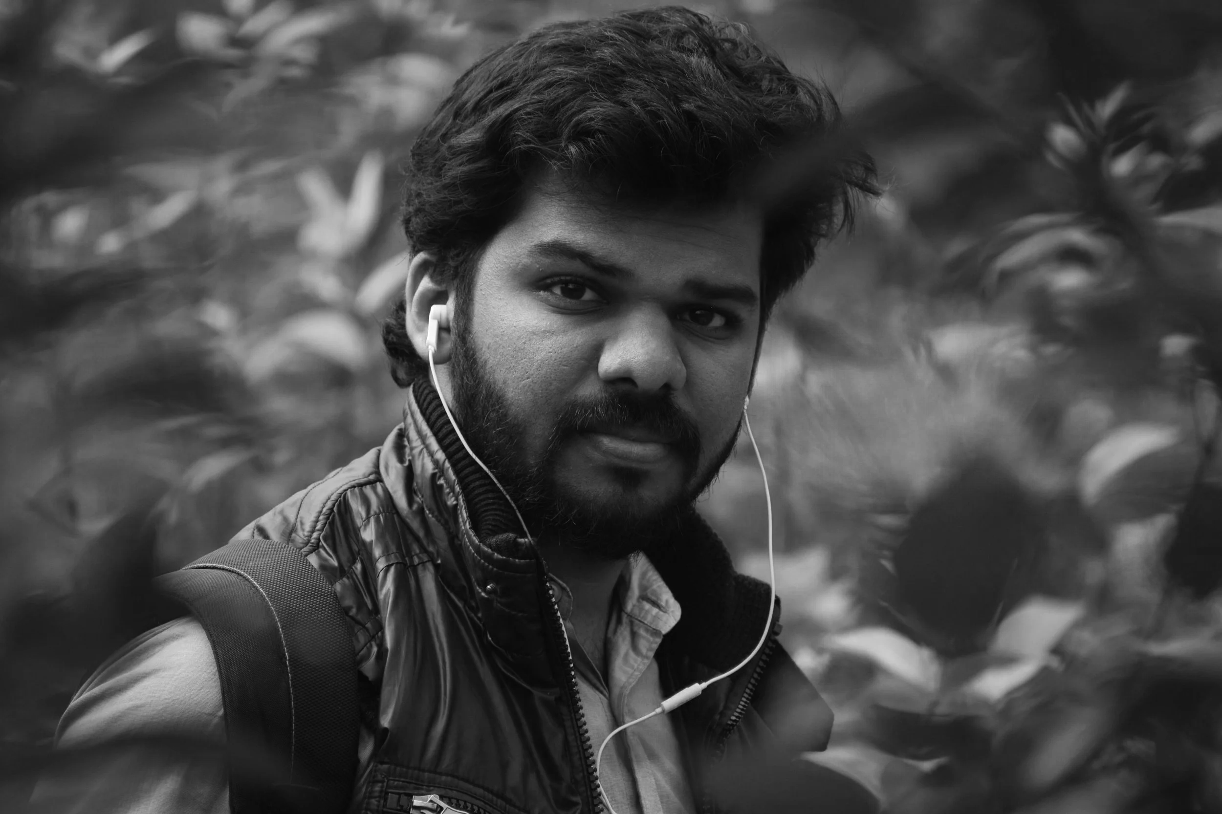 Black and white photo of a young man with a beard and dark hair, wearing earphones, a jacket, and a backpack, standing outdoors among blurred foliage.