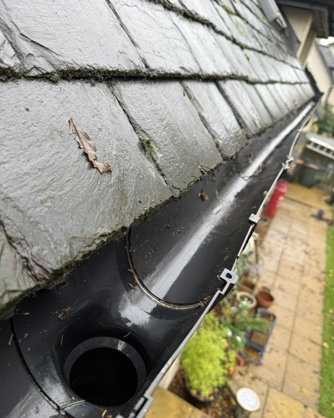 Close-up of a damaged gutter with moss and leaves, attached beneath a weathered gray shingled roof, with a garden and patio visible in the background.
