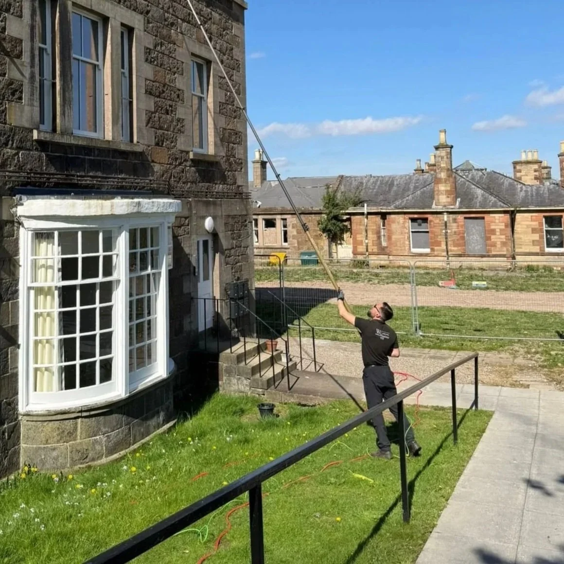 A man is cleaning a tall window cleaning tool attached to an extended pole outside a stone house on a clear, sunny day.