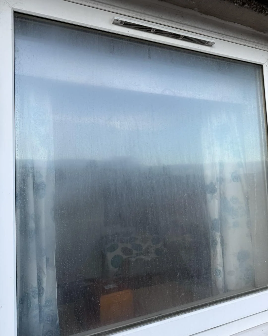 A window with a screen covered in condensation or fog, showing faint reflections of the outdoor scene and a partial interior view with patterned objects.