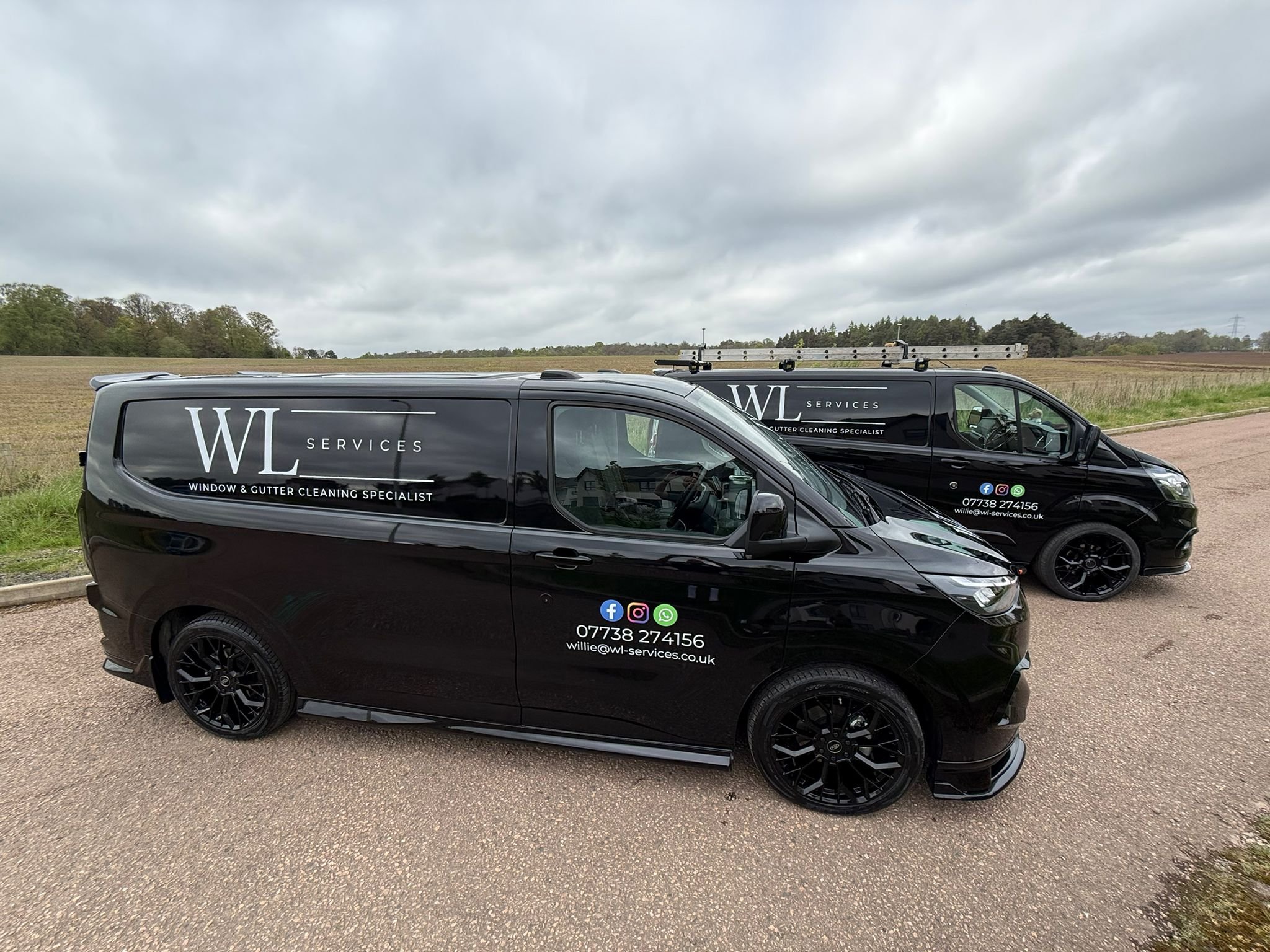 Two black vans with white lettering advertising a window and gutter cleaning service, parked on a paved area next to a grassy field under cloudy skies.