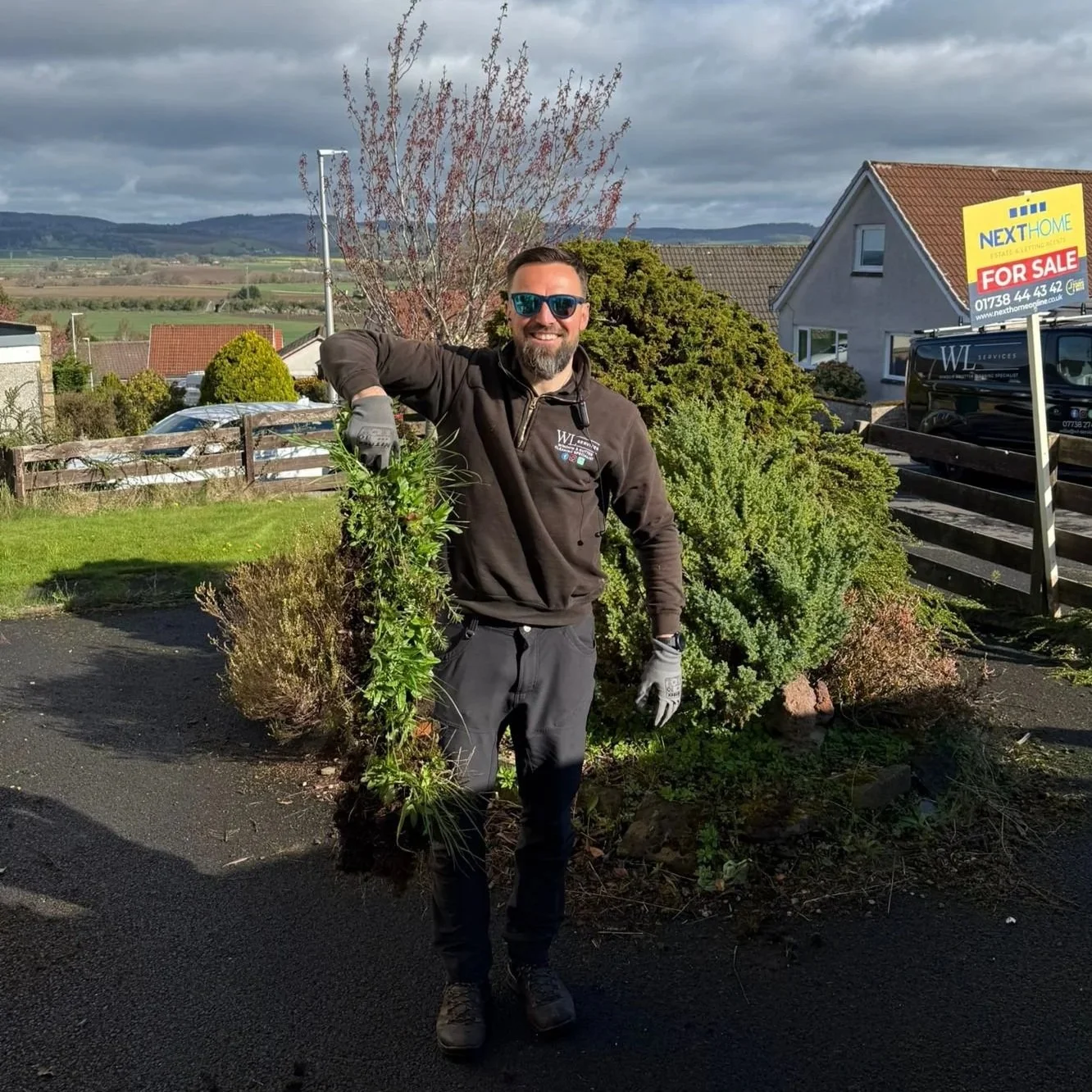A man with sunglasses and a beard wearing work gloves stands outdoors, holding a large green plant, with a landscaped yard, bushes, and houses in the background.
