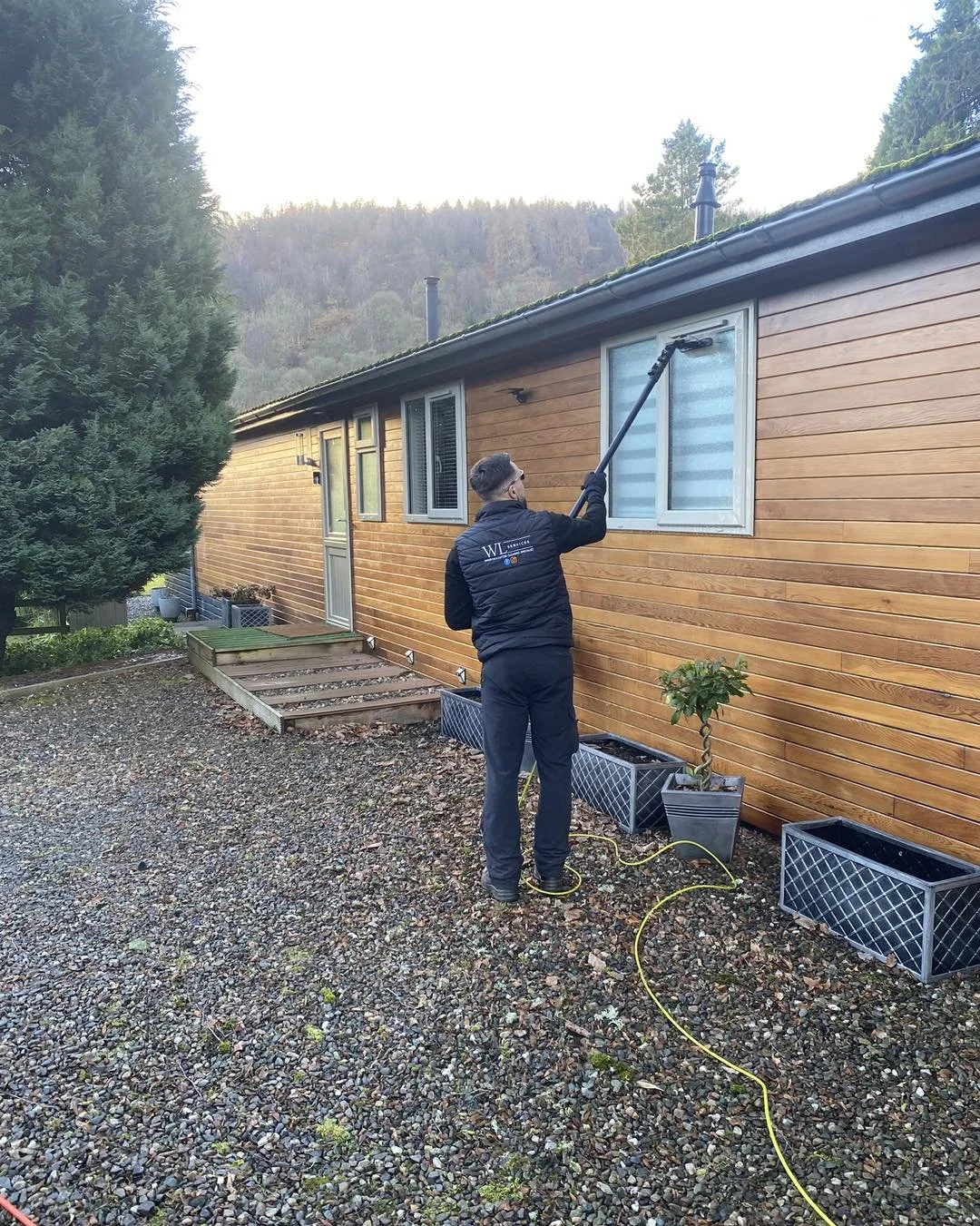 A man wearing a black jacket is pressure washing the exterior wall of a wooden house. The house has a door and windows, with plants in containers outside. There is a gravel yard and a large tree nearby, with a hill covered in trees in the background.