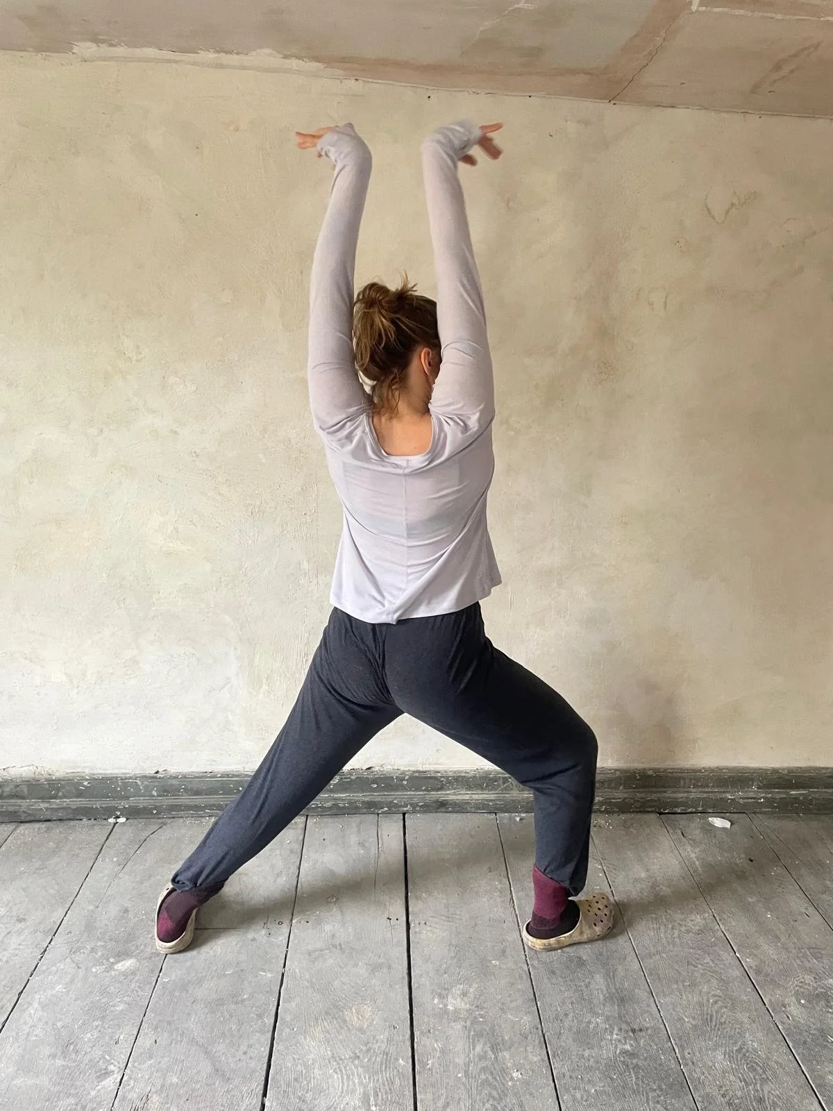 Person wearing gray sweatpants, a white long-sleeve shirt, and maroon and beige slippers, standing on a wooden floor with legs apart, arms raised overhead, in a stretching or yoga pose in a room with plain, light-colored walls.