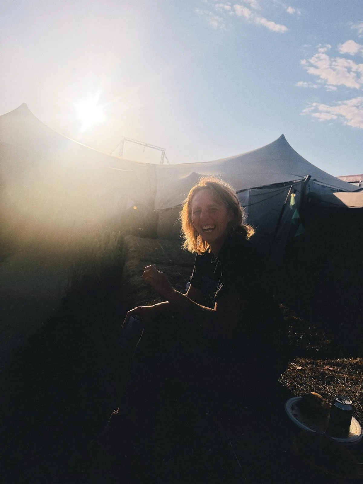 A woman with shoulder-length hair smiling and sitting outdoors at sunset, with tents and a partly cloudy sky in the background.
