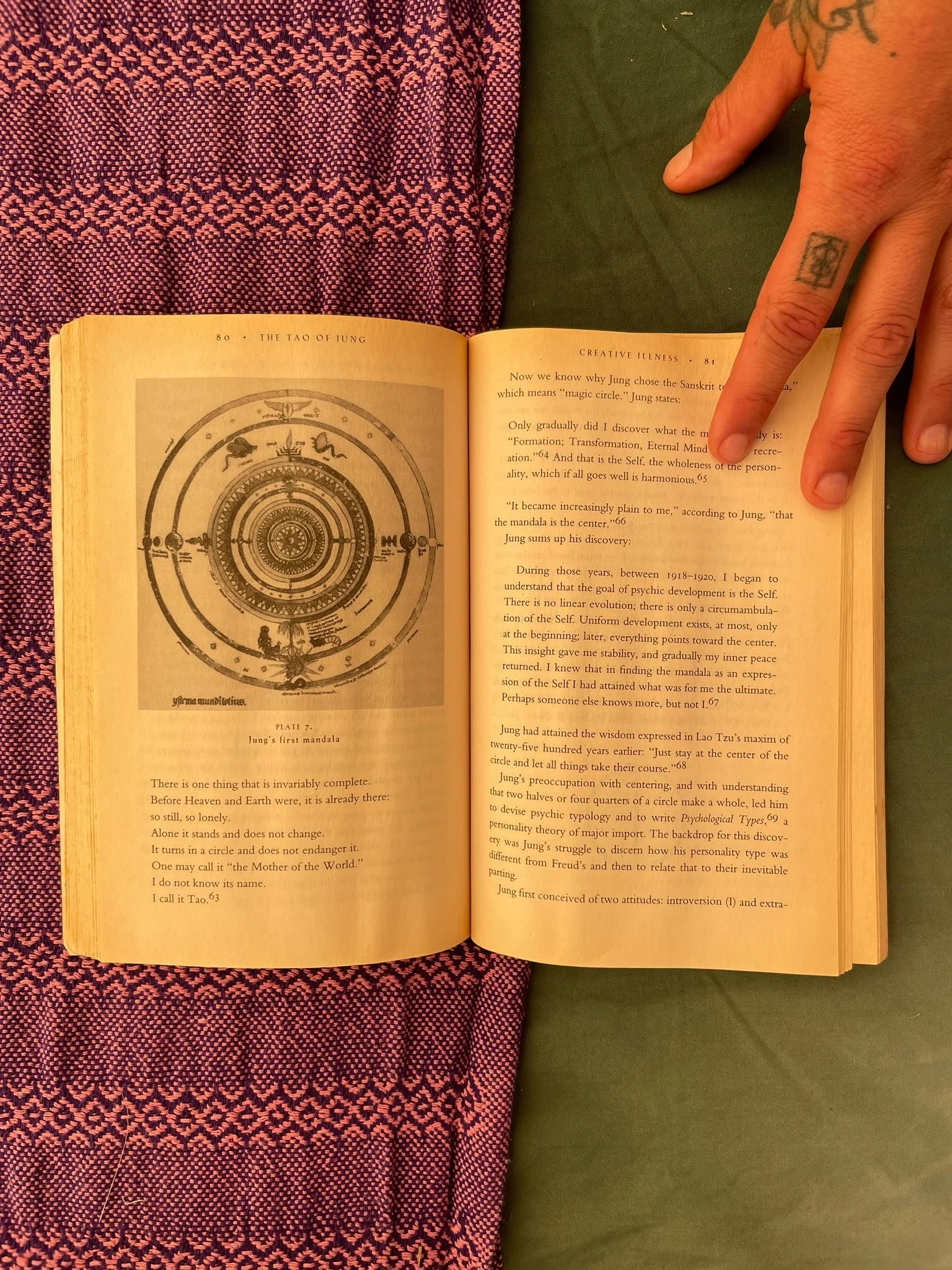 Open book showing a page with a diagram labeled "Jung's first mandala" and text discussing Jung's concept of mandalas and psychological development. The book is placed on a surface with a pink and purple patterned cloth on the left and a green cloth on the right, with a person's hand resting on the right side of the book.