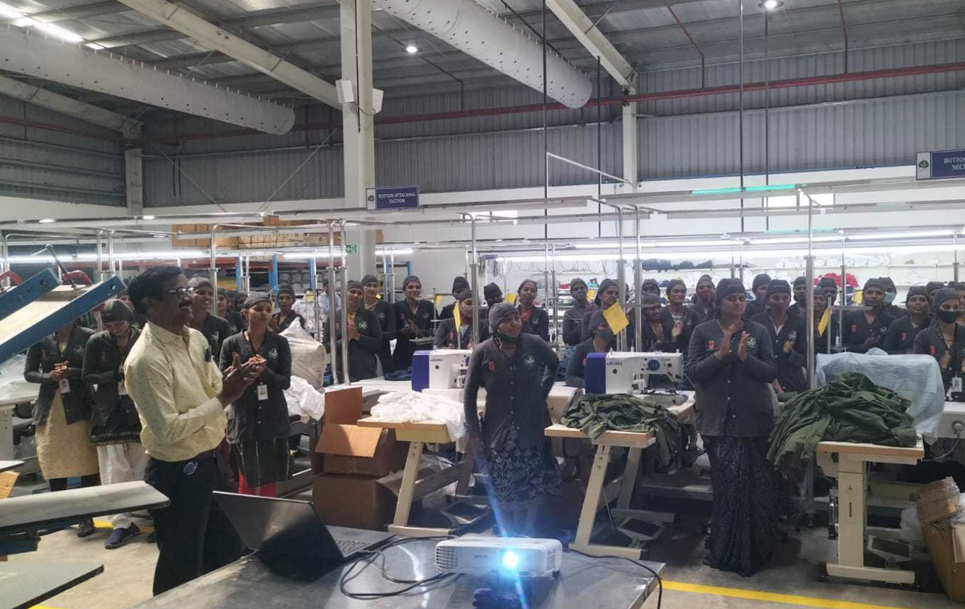 Workers in a factory or garment manufacturing facility listening to a speaker. The workers are wearing uniforms and some have masks. The factory has sewing machines and fabric on workstations.
