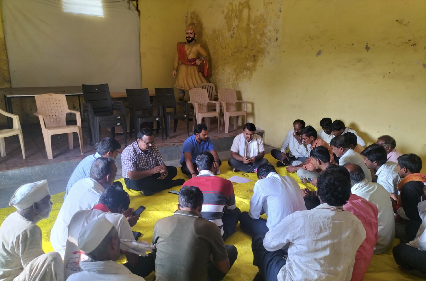 A group of men sitting on the floor in a room with yellow walls, some using their phones, while a statue of a man in traditional Indian attire stands against the wall in the background.