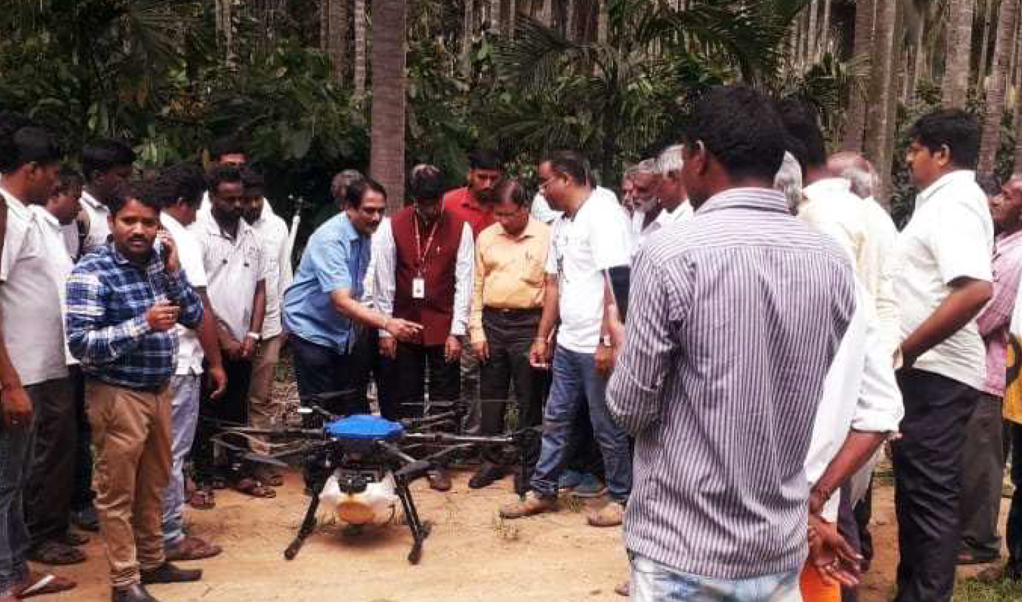 Group of men gathered outdoors around a drone, with tropical trees in the background.