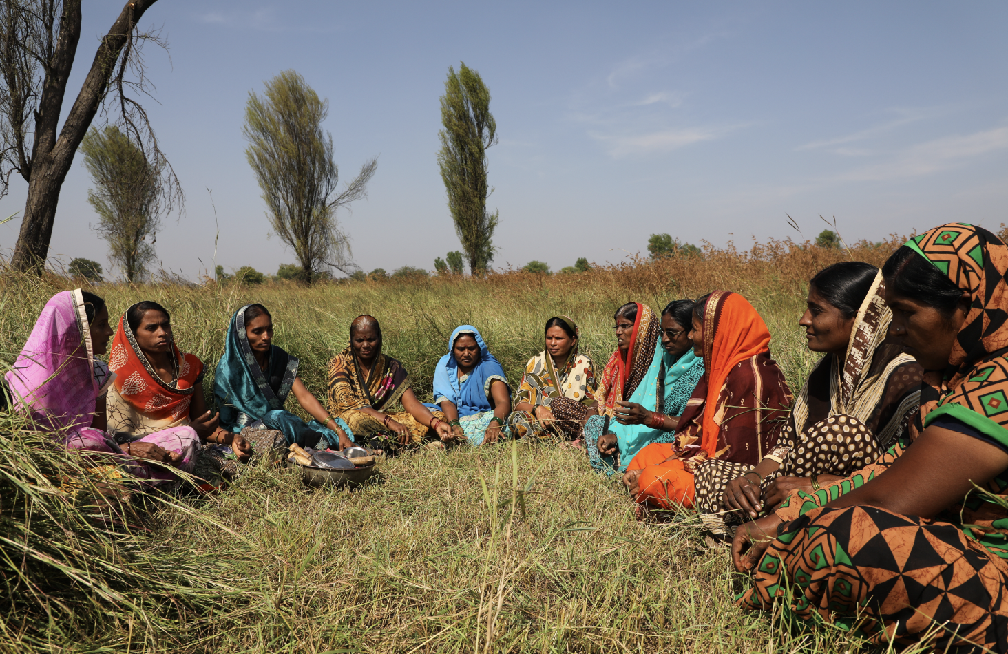 A group of women sitting on the grass in an open field, wearing colorful traditional clothing and headscarves.