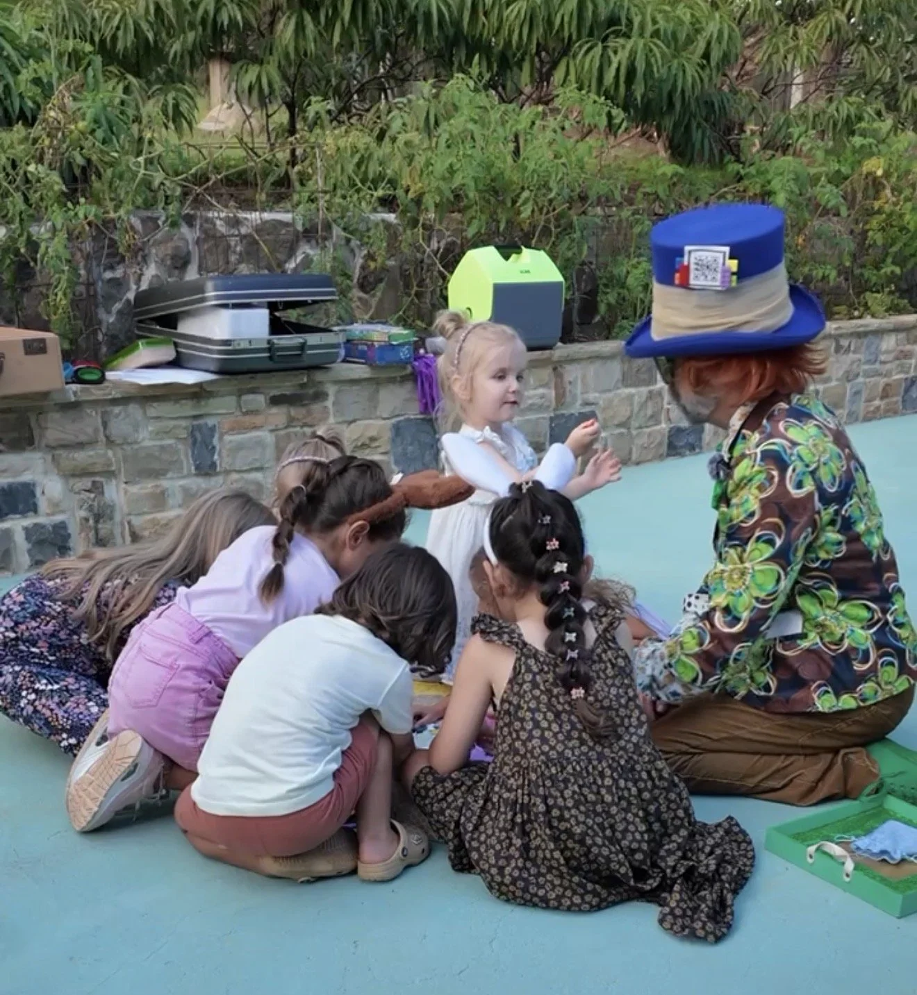 A person dressed as a wizard with a tall blue hat and patterned shirt is sitting on the floor with a group of children, some of whom are dressed in costumes, outdoors near a stone wall and plants. The children appear to be engaged in an activity, possibly a game or story session.