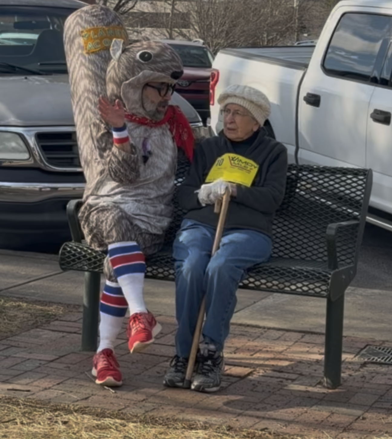 A person dressed in a squirrel costume sitting on a park bench talking to an elderly woman with glasses, a white knit hat, and gloves, holding a yellow flyer and a walking stick. The scene is outdoors with parked cars in the background.