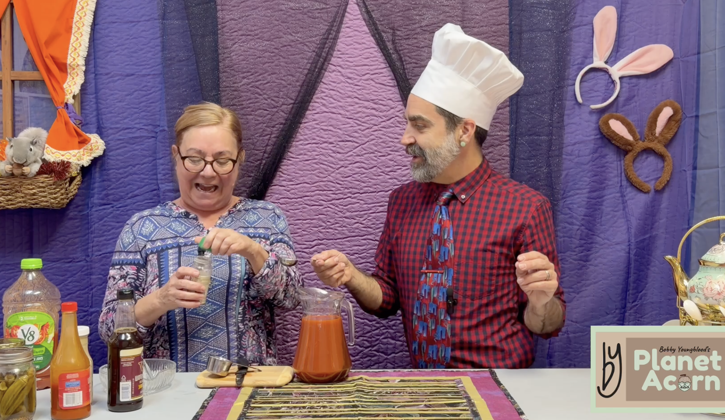 A woman with short blonde hair and glasses pouring a drink, and a man with a beard dressed as a chef with bunny ears on his head, standing at a table with various bottles and containers, in front of a purple quilted backdrop decorated with bunny ears.