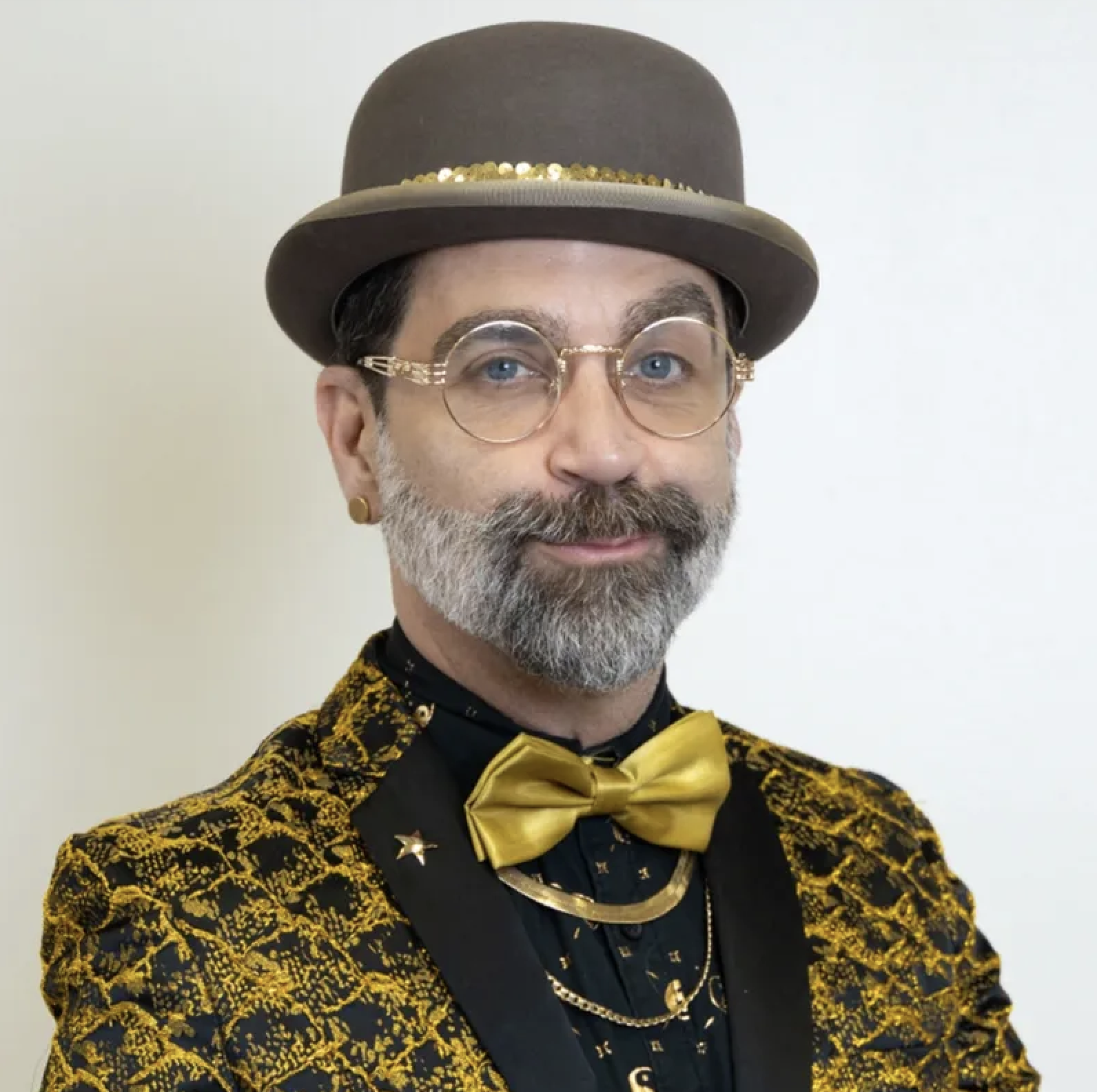 A man with a beard, glasses, and earrings wearing a brown hat with a band of gold sequins, a yellow and black patterned jacket, a black shirt with a gold bow tie, and gold accents, posing against a plain white background.