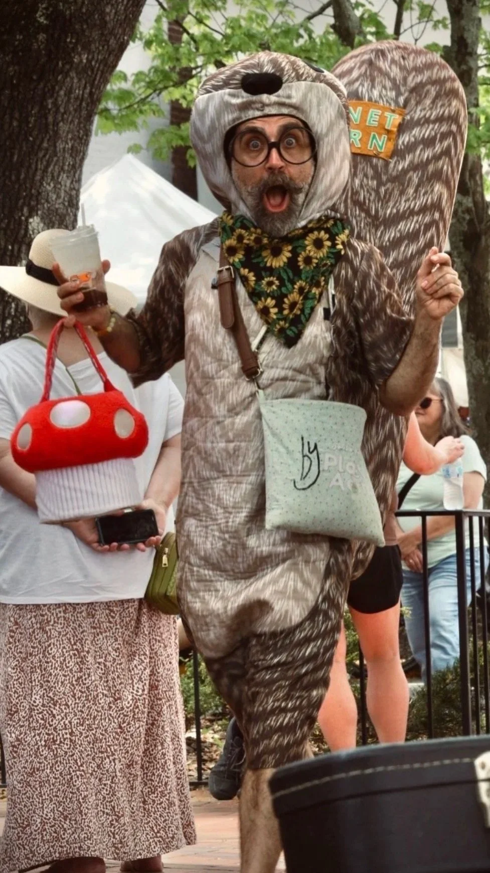 Man dressed as a bear with a surprised expression, wearing glasses, a bear costume, and a sunflower-patterned bandana, holding a drink, at an outdoor event.
