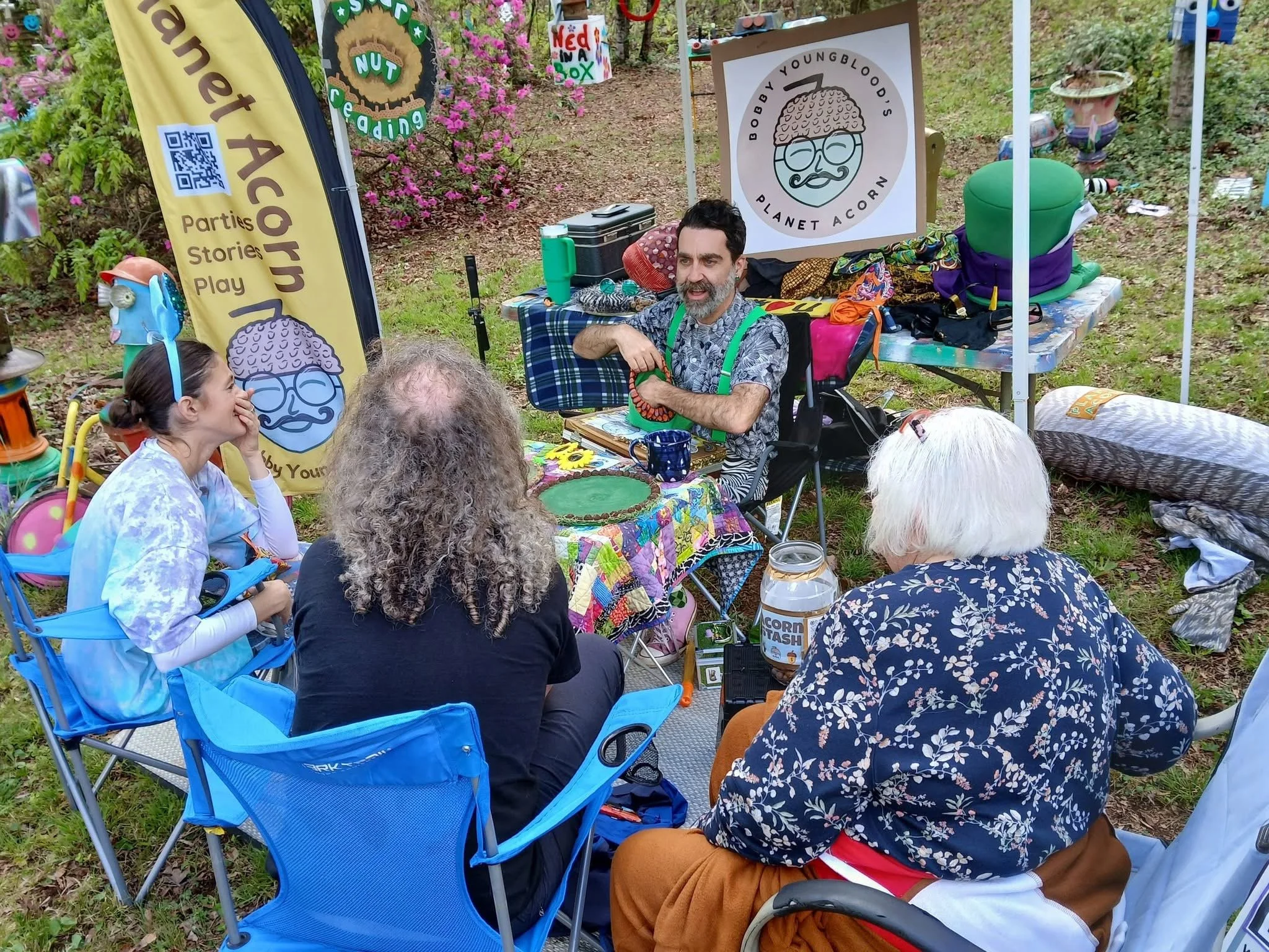Man sitting at a table with a group of people outdoors at a park, engaged in conversation, with colorful signs and plants in the background.