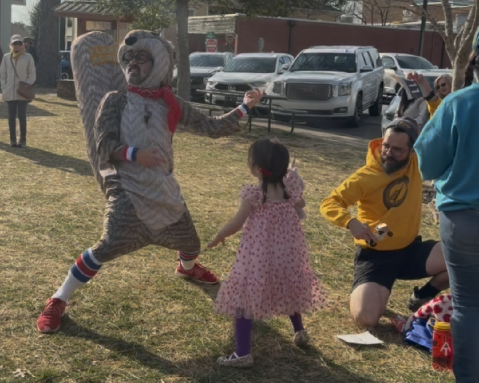 A man in a colorful playground costume, resembling a character or mascot, dancing and interacting with a young girl in a pink dress with red polka dots. Several adults are observing or seated nearby on a grassy field, with parked cars and buildings in the background.