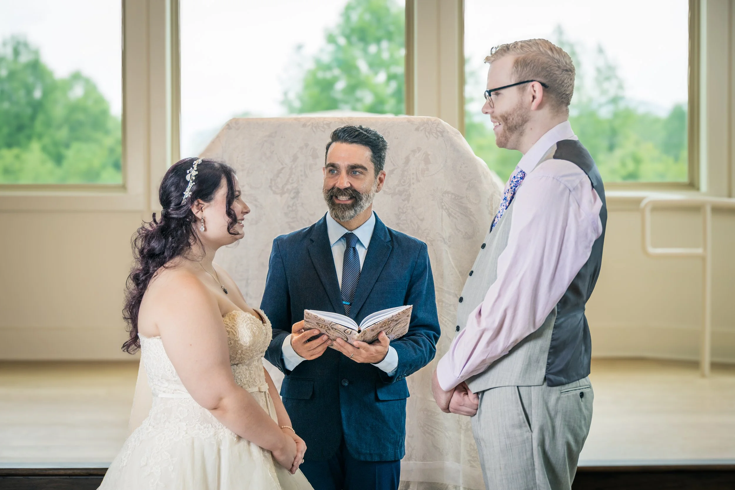 A bride and groom standing before an officiant during a wedding ceremony, exchanging vows in a bright room with windows and green trees outside.