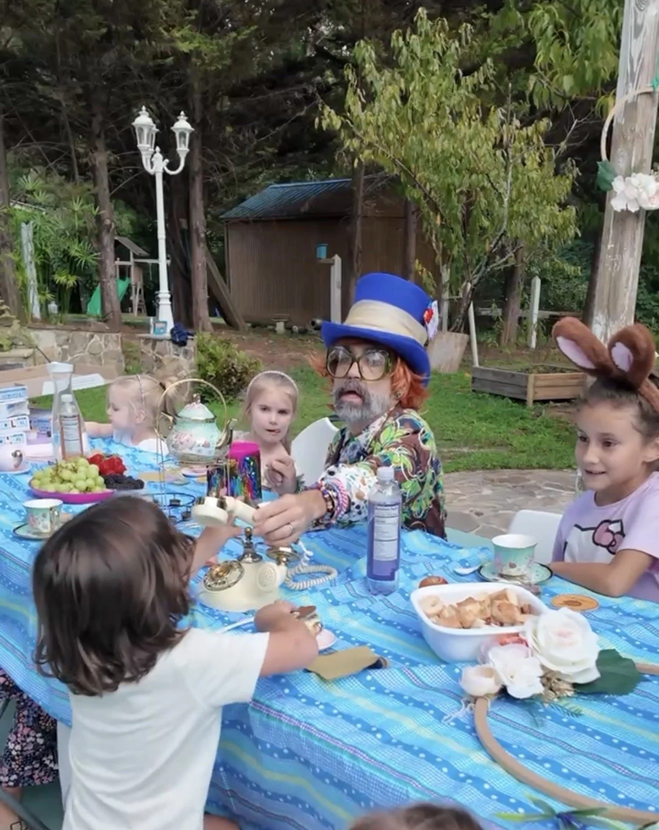Children and an adult wearing a colorful outfit and a blue top hat are gathered around a blue table outdoors with food and decorations, celebrating a party or holiday.