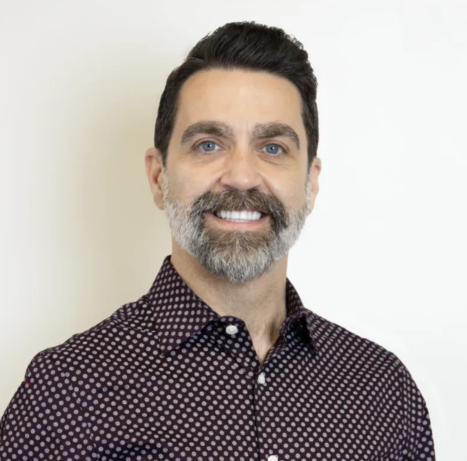 A smiling man with a beard and mustache, blue eyes, wearing a dark purple shirt with white dots, standing against a plain white background.
