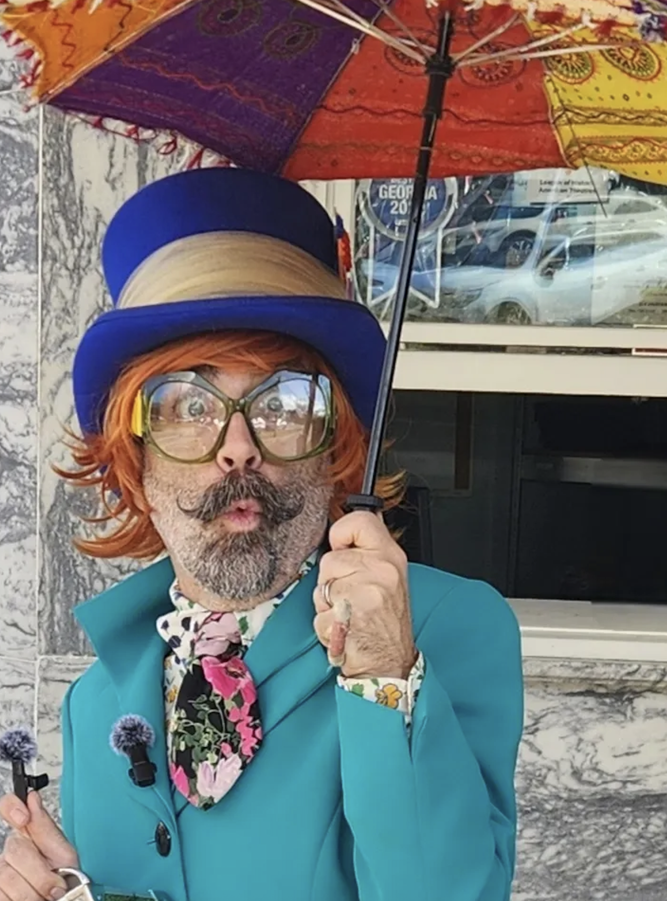A person dressed as a flamboyant character, wearing large glasses, an orange wig, a blue hat, a blue blazer, and a colorful scarf with floral patterns, holding a multicolored umbrella.