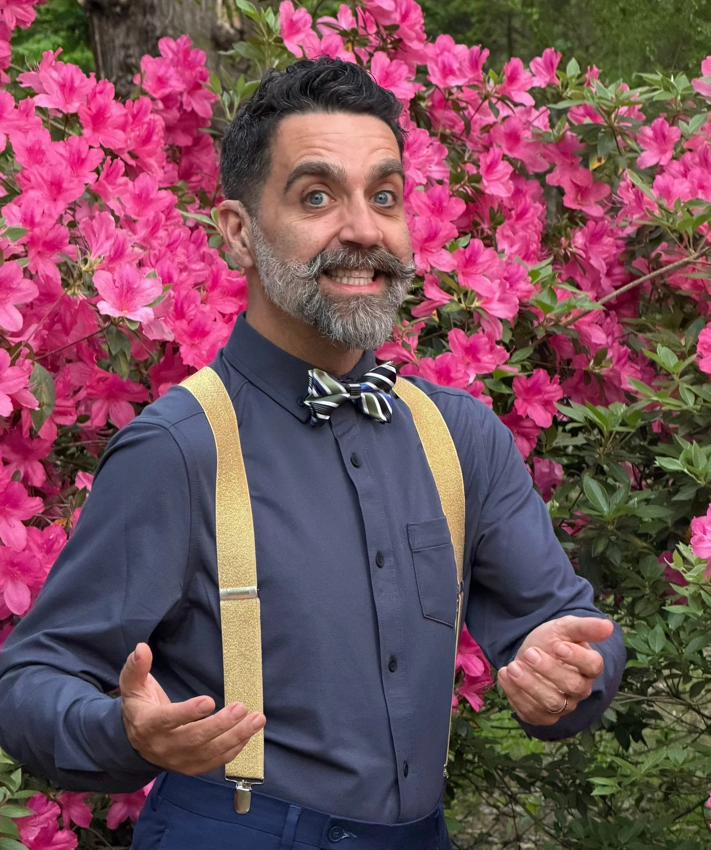 A man with dark, curly hair, a beard, and blue eyes smiling and gesturing with his hands in front of a large bush of pink flowers, possibly azaleas or rhododendrons.