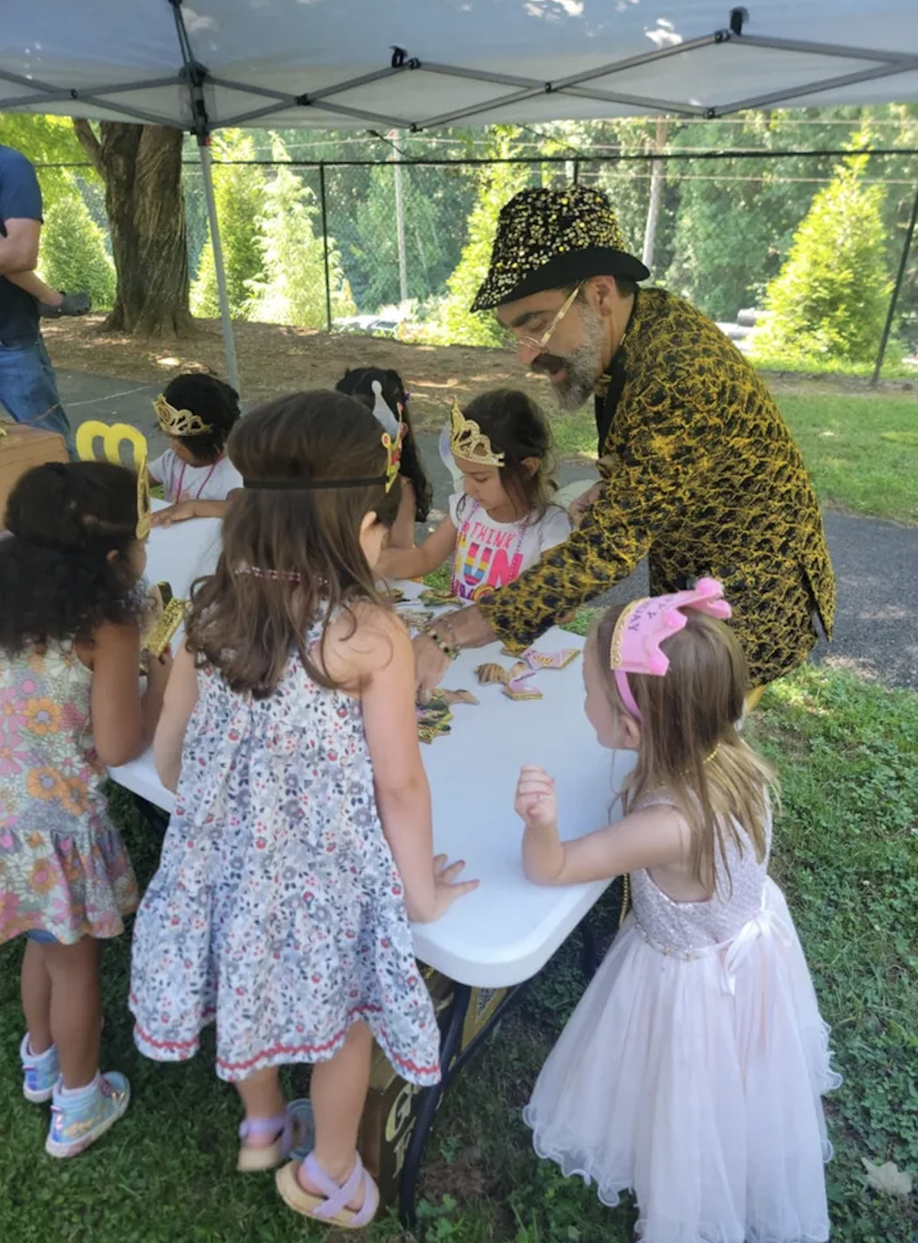 Children gathered around a table outdoors, wearing princess crowns, with an adult man in a leopard print jacket and a wizard hat assisting them. The setting is a park or backyard with trees and a fence in the background.