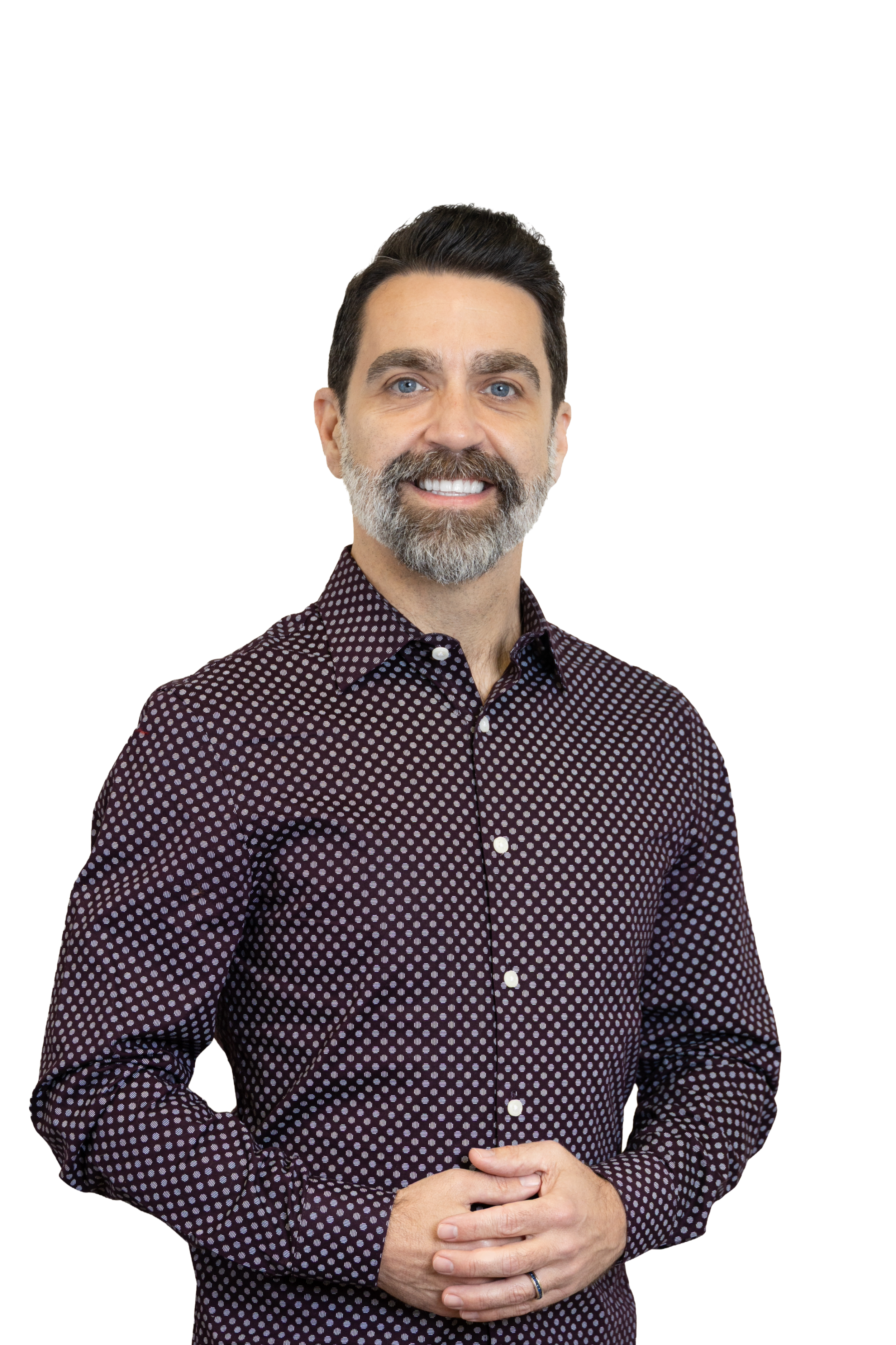 A middle-aged man with dark hair, a beard with grey, and blue eyes, smiling, wearing a dark patterned button-up shirt, standing with hands clasped.