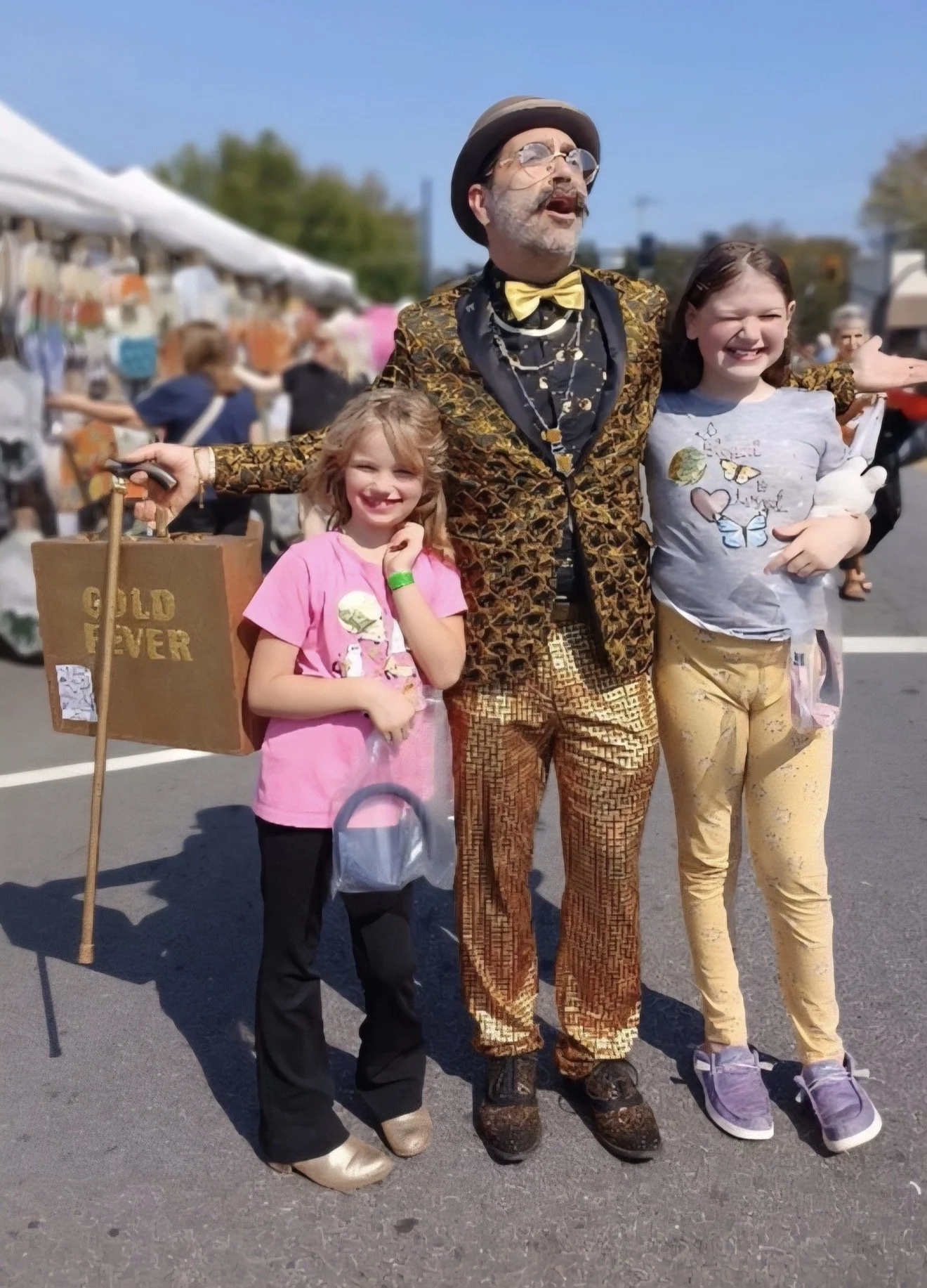 A street festival scene with three people in front of marketplace stalls. A man dressed in colorful, patterned suit, wearing a black hat, glasses, a yellow bowtie, and gold and black shoes. He has his arms around two young girls, both smiling. One girl in a pink shirt and black pants with beige shoes, and the other in a gray shirt with a butterfly design, yellow pants, and purple shoes. The girl in yellow holds a plush toy, and the girl in pink is holding a shopping bag. The background shows more festivalgoers and vendor tents on a sunny day.