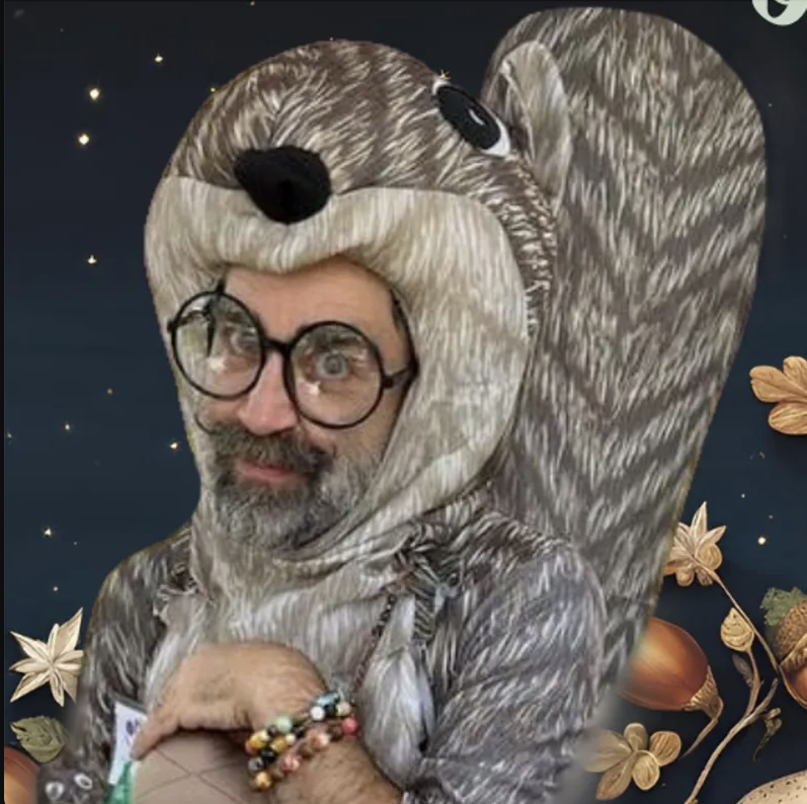 A person with glasses and a beard wearing a hoodie made to look like a fox, with a black nose and white facial markings. The background features autumn leaves and flowers with stars.