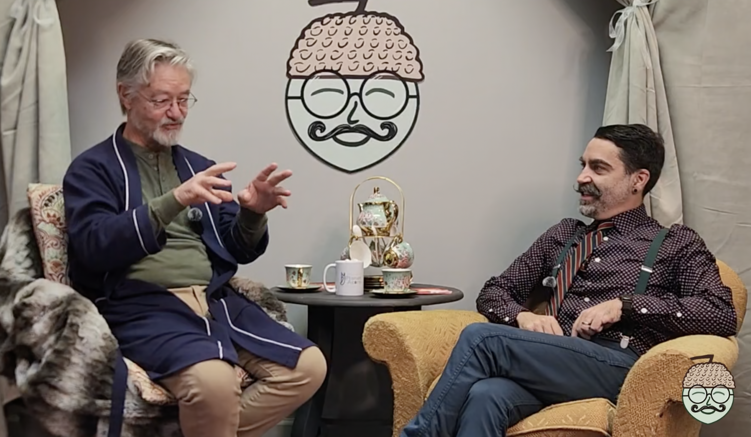 Two men sitting in a cozy room engaging in conversation, with tea cups and teapot on the table, and a large wall decoration of a stylized face with glasses, a mustache, and a brain on the wall behind them.