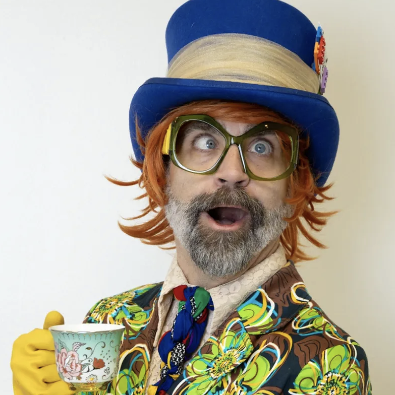 Man dressed in a colorful, patterned jacket and yellow gloves wearing large green glasses and a tall blue hat, with a surprised expression, holding a decorative cup. He has orange hair and a gray beard.