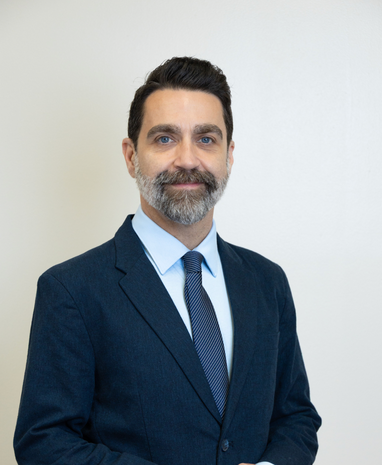 A professional portrait of a middle-aged man with dark hair, a beard and blue eyes, wearing a formal navy blue suit, light blue shirt, and a striped tie, standing against a plain light-colored background.