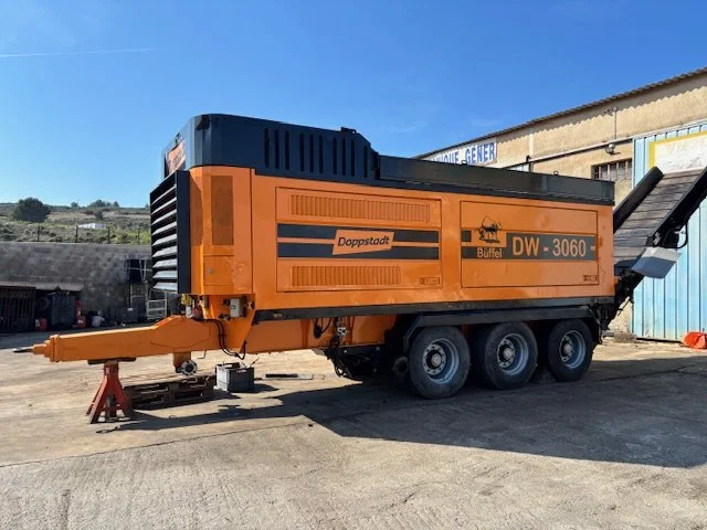 Un véhicule orange de type machine de construction ou de chantier avec des roues, stationné à l'extérieur sur une surface en béton, avec un bâtiment en arrière-plan.