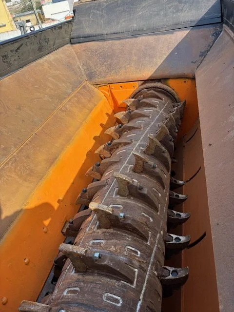 Une pièce mécanique, probablement une tête de coupe de machine agricole ou de construction, avec des dents métalliques fixées sur une tige centrale, dans un compartiment orange.