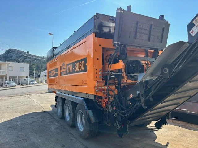 Un véhicule de construction orange équipé d'une machine pour la pose de pavés ou d'autres matériaux, stationné sur une route.